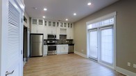 This single-floor space offers a thoughtfully designed open-concept layout that seamlessly integrates the kitchen and living areas. The kitchen is a standout feature, showcasing floor-to-ceiling white cabinetry with frosted glass upper panels, dark granite countertops, and a mosaic tile backsplash that adds texture and visual interest. A full suite of stainless steel appliances — including a refrigerator, range, microwave, and dishwasher — provides both functionality and a sleek, contemporary aesthetic. A wine rack detail accents the upper cabinetry, adding a refined touch. The open living area is finished with light hardwood flooring that runs throughout, creating a warm and cohesive atmosphere. Recessed lighting illuminates the space evenly, complementing the natural light that pours in through a large floor-to-ceiling window with sliding door access. The neutral, warm-toned walls enhance the bright and airy feel of the interior. The overall ambiance is clean, modern, and welcoming — ideal for both everyday living and entertaining.