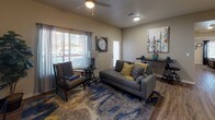 This single-story residence offers a cohesive, modern design highlighted by wood-look flooring that flows consistently throughout the space. The living room serves as a central gathering area, featuring a ceiling fan with light fixture, large windows dressed with sheer curtains and blinds that allow natural light to fill the room, and ample space for both a sofa arrangement and dedicated seating area.

The kitchen is equipped with light wood cabinetry, marble-patterned countertops, and stainless steel appliances including a full-size refrigerator and range, providing functional workspace for meal preparation.

The primary bedroom is generously sized, accommodating a full bedroom suite, and is outfitted with a ceiling fan, wood-look flooring, and a large window. It connects to a spacious bathroom featuring light wood vanity cabinetry, marble-style countertops, wood-look flooring, and direct access to a walk-in closet with built-in shelving. The bathroom also includes a soaking tub with floor-to-ceiling neutral-toned tile surround.

An additional bedroom with a built-in closet offers flexibility as a guest room or home office. The floor plan, as viewed in the dollhouse render, reflects a well-organized layout with clear separation between living and sleeping areas.