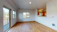 This single-floor unit features a straightforward, functional layout with a clean, modern aesthetic throughout. Light-toned hardwood-style flooring runs through the main living areas, while carpeting provides comfort in the bedrooms. The living area is well-proportioned, with multiple windows offering views of the surrounding outdoor scenery, including mature trees, and provides direct access to a private balcony through a glass door flanked by windows. The kitchen is positioned adjacent to the living space, connected via a pass-through counter opening that maintains a sense of openness. It is equipped with warm-toned wood cabinetry, white countertops, a stainless steel undermount sink, and a window above that brings in natural light and outdoor views. A central hallway with wood-trimmed doorways leads to the bedrooms and bathroom. The bedrooms feature neutral carpet, white walls, and windows that bring in natural light. The bathroom includes a dual-sink vanity with white countertops and wood cabinetry, framed mirrors, hexagonal tile flooring, and a full soaking tub with a tiled shower surround. A utility closet housing the water heater is also accessible from the bathroom area.