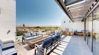 Located at 525 N 7th St in San Jose, California, this rooftop amenity space offers a well-appointed outdoor retreat designed for relaxation and socializing. The expansive rooftop terrace features a contemporary aesthetic defined by clean white stucco walls, large-format tile flooring, and sleek glass panel railings that preserve unobstructed panoramic views of the surrounding cityscape and the distant mountain range beyond.

The main lounge area is anchored by a substantial rectangular fire pit, surrounded by generously cushioned outdoor sofas and armchairs in a cool blue palette with coordinating wood-framed furniture. A separate, more intimate courtyard-style nook provides a semi-sheltered seating zone with built-in wooden bench seating, colorful accent cushions, and bistro-style tables and chairs, ideal for casual gatherings.

Floor-to-ceiling glass walls and sliding doors connect the outdoor terrace to an adjacent interior space, allowing abundant natural light to flow freely between the two areas. An overhead pergola structure provides partial shade to portions of the deck, enhancing comfort throughout the day. Together, these elements create a vibrant, modern outdoor environment well-suited for both quiet retreat and community engagement.