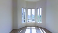 This single-floor space makes a strong first impression with its airy, open layout and refined modern aesthetic. The most commanding feature is a sweeping bay window arrangement, composed of multiple tall, white-framed windows that wrap around a corner of the main room. These expansive windows flood the interior with abundant natural light, casting bright, geometric patterns across the warm hardwood flooring below.

The ceilings rise impressively above, amplifying the sense of openness and volume within the space. Walls are finished in a crisp, neutral white, creating a clean and versatile backdrop that enhances the brightness already introduced by the generous glazing.

The hardwood floors offer a warm, natural contrast to the cool white walls and window frames, lending the space a balanced and welcoming ambiance. From the windows, views extend outward to surrounding buildings and lush green treetops, connecting the interior to its urban surroundings.

The overall atmosphere is bright, modern, and serene, with an emphasis on natural light, clean lines, and an uncluttered sense of space. The thoughtful architectural details — particularly the multi-panel bay windows and elevated ceilings — give the space a distinctive character well-suited to a variety of uses.