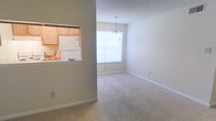 This single-floor residence offers a bright and functional layout designed for comfortable everyday living. The dining area is a welcoming focal point, featuring soft wall-to-wall carpeting underfoot and a large picture window that allows abundant natural light to pour into the space. An elegant chandelier hangs above, adding a refined touch to the room's ambiance.

A distinctive pass-through window connects the dining area directly to the kitchen, creating an open and interactive feel between the two spaces. The kitchen is equipped with light wood cabinetry and a white refrigerator, conveying a clean and practical aesthetic. The pass-through also serves as a convenient counter space, ideal for casual serving or entertaining.

The interior palette throughout is neutral and understated, with light-toned walls and white trim that contribute to a fresh, airy atmosphere. The consistent use of soft carpeting and clean lines gives the space a cohesive and approachable character. Overall, this residence combines practical design with inviting details, making it a comfortable and well-lit living environment suited to a variety of lifestyles.