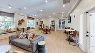 Located at 13625 Haven Ridge Ln in Charlotte, NC, this single-floor community clubhouse offers a thoughtfully designed gathering space that blends comfort with modern style. The open-plan layout is anchored by light hardwood flooring throughout, with crown molding and recessed lighting that create a polished, welcoming atmosphere.

The main lounge area features a generous sectional sofa, coordinated accent chairs, and a wall-mounted flat-screen television set against a bold navy accent wall, providing a relaxed setting for socializing or entertainment. Adjacent to the lounge, a round dining table with curved mid-century style chairs offers an inviting spot for casual meals or small group gatherings.

A dedicated co-working zone lines one side of the room, equipped with individual workstations, desktop computers, and bistro-height tables with bar seating — ideal for residents seeking a productive environment. Built-in window benches flanking large windows flood this area with natural light and offer comfortable perch seating.

A stylish refreshment nook features deep navy cabinetry with brushed gold hardware, white quartz countertops, a geometric patterned tile backsplash, and a water dispenser accompanied by a curated coffee and tea station. French doors open to an outdoor space, further enhancing the bright, airy feel of the entire clubhouse.