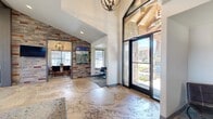 This well-appointed single-story community or leasing facility offers generous square footage across four distinct functional areas, thoughtfully designed to serve both social and professional needs. The grand entrance immediately impresses with soaring ceilings, floor-to-ceiling black-framed glass doors, and a striking combination of natural stone cladding and reclaimed wood accent walls that establish the space's modern rustic aesthetic. Polished concrete flooring runs throughout, adding an industrial yet refined touch. The open lounge area is anchored by a prominent service counter with navy cabinetry and bar-height seating, complemented by vaulted, beam-accented ceilings, recessed lighting, and elegant chandelier fixtures. Comfortable seating arrangements, layered area rugs, and curated artwork create a warm and inviting atmosphere. Adjacent rooms include dedicated conference spaces featuring dark stone-topped tables, mid-century modern chairs, oversized windows with roller shades, and pendant globe chandeliers that provide intimate meeting environments bathed in natural light. A private office, finished with bold blue walls and eclectic décor, offers a personalized workspace. Throughout, large windows frame views of the well-maintained exterior grounds, ensuring the interior remains bright and connected to the outdoors.