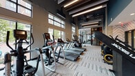 This expansive fitness center is designed with a contemporary aesthetic, combining high-performance functionality with thoughtful interior design. The space is defined by its impressive double-height ceilings fitted with dark exposed beams and sleek linear pendant lighting, creating an energetic yet polished atmosphere. Large floor-to-ceiling windows line multiple walls, flooding the interior with natural light and offering open views of the surrounding streetscape.

The cardio zone is generously equipped with multiple treadmills, ellipticals, stair climbers, and stationary bikes, all arranged to take advantage of the natural light and outdoor views. Adjacent to this area, a comprehensive strength training section features cable machines, benches, and a full rack of Matrix-branded free weights and resistance equipment.

A dedicated functional training zone showcases a well-organized rack system stocked with kettlebells, medicine balls, stability balls, TRX suspension trainers, battle ropes, and a heavy punching bag, set against a striking chevron-patterned accent wall. A water station is conveniently positioned nearby.

The fitness center also incorporates a comfortable lounge area with vibrant red chairs and a round accent table, offering a place to rest between workouts. Patterned carpet tiles throughout the space tie the design together cohesively.