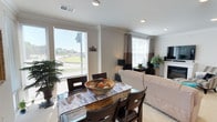 This single-floor space features an open-plan layout that seamlessly connects the dining and living areas, creating a cohesive and functional environment. Crown molding along the ceiling adds a refined architectural detail throughout the space, complementing the neutral, warm-toned walls.

The dining area is anchored by a dark wood table with a marble-patterned glass top, surrounded by matching dark wood chairs. A decorative wooden bowl serves as a centerpiece, adding a natural, earthy touch. Large windows with horizontal blinds flood this area with abundant natural light while offering views of the surrounding outdoor landscape.

The adjacent living area features a comfortable slipcovered sofa and is enhanced by tall curtained windows that continue the bright and airy atmosphere. A television setup and a white fireplace mantel provide functional and aesthetic focal points on the far wall. Lush potted plants are thoughtfully placed throughout, introducing vibrant greenery that enlivens the neutral palette.

Recessed lighting complements the natural light, ensuring the space remains well-lit throughout the day. The overall ambiance is clean, welcoming, and casually elegant.