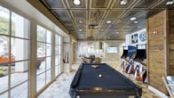 THE HQ's entertainment lounge is a thoughtfully designed recreational space that combines classic and contemporary elements. The room's most striking architectural feature is its ornate tin-style ceiling, finished in dark metallic tones with an intricate geometric pattern and embedded recessed lighting that casts a warm, sophisticated glow throughout the space.

The flooring features a decorative geometric tile pattern that adds visual interest and complements the room's upscale aesthetic. Floor-to-ceiling glass doors and large windows along one wall flood the space with natural light while offering views of the surrounding outdoor greenery.

A full-size billiards table with a dark felt surface serves as a central focal point, suspended above by a cylindrical pendant light fixture. Toward the back of the room, a dedicated dining or gaming table with surrounding chairs provides a secondary gathering area, accompanied by a wall-mounted flat-screen television. To the right, a vibrant arcade cabinet adds a playful, retro touch to the lounge's overall character.

The combination of the ornate ceiling, natural light, tile floors, and varied recreational amenities creates an engaging, well-appointed space ideal for social gatherings and leisure activities.