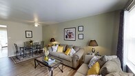This two-floor residence offers a thoughtfully arranged living environment with a welcoming and comfortable atmosphere throughout. The main living area is finished with light wood-style flooring that extends seamlessly across the space, complemented by soft sage-green walls that lend a calm, neutral backdrop. A ceiling fan overhead provides both function and a casual aesthetic, while warm table lamps add layered lighting to the room.

The living room is generously proportioned, centered around plush, neutral-toned sofas accented with yellow and patterned throw pillows that add a pop of color and personality. A glass-top coffee table anchors the seating arrangement, and framed artwork decorates the walls, adding visual interest and character to the space.

An open sightline connects the living area to a dedicated dining space, where a dark-framed table and matching chairs create an intimate setting for meals. The open-plan layout enhances the sense of spaciousness and promotes easy movement between areas.

Spanning two floors, this residence provides ample room across multiple levels, offering separation between living and private spaces. The overall ambiance is warm and livable, with a cohesive design palette of neutral tones, natural textures, and tasteful décor accents that come together to create an inviting home environment.