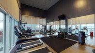 This fitness center offers a comprehensive workout environment designed with both functionality and aesthetics in mind. The space is characterized by soaring ceilings and dark, sophisticated wall tones that create a focused, energetic atmosphere. Floor-to-ceiling windows line the perimeter, flooding the space with natural light and providing expansive city skyline views, enhancing the workout experience throughout.

The cardio zone features a row of True-brand treadmills positioned directly in front of the windows, alongside elliptical trainers and stationary cycling bikes, all equipped with individual screens. A dedicated strength training area includes a multi-function cable machine, a dumbbell rack with a full range of free weights, and flat benches for varied exercises. A wall-mounted speed bag and boxing station occupy a separate windowed corner, offering an additional training option.

Full-length mirrors run along one wall, amplifying the sense of space and allowing users to monitor their form. Rubber floor mats define the free-weight and stretching zones. A wall-mounted flat-screen television is positioned for visibility across the main floor. The cork-style flooring throughout adds durability and comfort underfoot, completing a well-appointed space suited for a wide range of fitness routines.