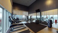 This fitness center offers a comprehensive workout environment designed with both functionality and aesthetics in mind. The space is characterized by soaring ceilings and dark, sophisticated wall tones that create a focused, energetic atmosphere. Floor-to-ceiling windows line the perimeter, flooding the space with natural light and providing expansive city skyline views, enhancing the workout experience throughout.

The cardio zone features a row of True-brand treadmills positioned directly in front of the windows, alongside elliptical trainers and stationary cycling bikes, all equipped with individual screens. A dedicated strength training area includes a multi-function cable machine, a dumbbell rack with a full range of free weights, and flat benches for varied exercises. A wall-mounted speed bag and boxing station occupy a separate windowed corner, offering an additional training option.

Full-length mirrors run along one wall, amplifying the sense of space and allowing users to monitor their form. Rubber floor mats define the free-weight and stretching zones. A wall-mounted flat-screen television is positioned for visibility across the main floor. The cork-style flooring throughout adds durability and comfort underfoot, completing a well-appointed space suited for a wide range of fitness routines.
