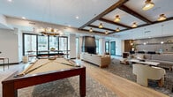 This expansive single-floor amenity lounge is thoughtfully designed to blend sophistication with a relaxed, social atmosphere. The space is anchored by rich, wide-plank hardwood flooring that flows throughout the main lounge area, complemented by decorative exposed wood ceiling beams that add warmth and architectural character. Recessed lighting and a mix of sculptural pendant fixtures illuminate the space with a warm, inviting glow.

The centerpiece of the lounge is a full-size billiards table positioned beneath a striking chandelier, with a shuffleboard table nearby for additional recreation. Large windows allow natural light to fill the room while offering views of the surrounding streetscape. A sleek, dark fireplace serves as a dramatic focal point in the seating area, which features neutral-toned sofas arranged for comfortable gathering.

The wet bar area is a standout highlight, showcasing an oversized marble-slab island with gold veining, leather bar seating, warm wood cabinetry, and an intricate geometric tile floor with gold accents. Pendant lighting above the island adds an elegant finishing touch.

Built-in shelving with dark-painted interiors provides display and storage space, while a decorative chain-link partition near the entry adds an artistic, industrial edge. The overall ambiance is modern yet approachable, ideal for entertaining.