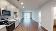 This well-appointed single-floor residence offers a thoughtfully designed open-concept layout that seamlessly connects the kitchen and living room, creating a bright and functional living space. Rich wood-style flooring runs throughout the main areas, complementing the clean, contemporary aesthetic defined by crisp white walls and recessed lighting.

The kitchen is a standout feature, outfitted with stainless steel appliances — including a French door refrigerator, electric range, and over-the-range microwave — paired with light wood-grain cabinetry, white quartz countertops, and a subtle gray subway tile backsplash. The living room is bathed in natural light through large sliding glass doors fitted with vertical blinds, opening to an outdoor view.

The generously sized primary bedroom features plush carpeting, multiple windows with exterior views, and direct access to a connected room, offering a comfortable private retreat. Consistent with the modern design, the bedroom maintains neutral tones and clean lines throughout.

A dedicated laundry area houses a stacked washer and dryer unit and includes overhead shelving for additional storage. The entryway provides a practical transition point between the kitchen and living areas, completing this efficiently organized and move-in-ready residence.