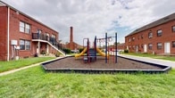 Located at 7003 Dunmanway in Dundalk, Maryland, this two-story residential community is built with classic red brick construction, reflecting a durable and traditional architectural style. The multi-unit row-style buildings are arranged throughout the property, offering a cohesive neighborhood layout with open green spaces running along the front and rear of each building. Each unit features individual entry doors with front stoops, and select units on the upper level are equipped with wooden balconies accessible via exterior staircases, providing private outdoor space with views of the surrounding grounds. Windows are generously distributed across both floors, allowing natural light into the interiors. The community grounds include well-maintained grassy areas that create an open, breathable atmosphere between the buildings. A dedicated children's playground with colorful multi-activity equipment, slides, and a mulch safety surface serves as a central outdoor amenity for residents. Internal roadways and walkways connect the various sections of the property. Mature trees and landscaped shrubs are present throughout, adding greenery to the environment. The overall layout promotes a close-knit community feel, with shared outdoor spaces and pedestrian-friendly pathways running throughout the development.