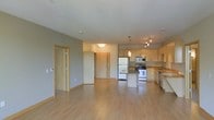 This single-floor space is designed with an open-concept layout that integrates the kitchen and living areas into one cohesive, functional environment. Light hardwood flooring extends throughout, contributing to a bright and airy atmosphere. The kitchen is well-appointed with light maple cabinetry, stainless steel appliances including a refrigerator, electric range, and built-in microwave, as well as a central island with a dark countertop that provides additional workspace and seating potential. A pendant light fixture above the island adds a focused design element, complemented by a track lighting system overhead. The countertops along the perimeter feature a sink and ample prep space. The open living area offers generous room for furnishing arrangements, with neutral gray walls providing a versatile backdrop. Two interior doors visible from the main space lead to additional rooms, suggesting a practical and well-organized floor plan. Recessed lighting and a flush-mount ceiling fixture ensure consistent illumination throughout. The overall aesthetic is clean, contemporary, and functional, with warm wood tones balancing the neutral palette.