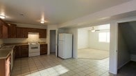 This two-floor space offers a functional and open layout designed for comfortable everyday living. The kitchen is equipped with dark wood cabinetry, a white electric range, and a full-size refrigerator, and is finished with light ceramic tile flooring that reflects natural light throughout the space. The kitchen flows seamlessly into the adjacent living area, which transitions to plush carpeted flooring, creating a clear distinction between the two zones while maintaining an open, connected feel. The living area is equipped with a ceiling fan, contributing to air circulation and comfort, and features a window that allows natural light to brighten the room. A doorway visible from the kitchen leads to additional rooms or utilities, suggesting practical access to further spaces within the unit. The neutral color palette of white walls and light tones throughout gives the space a clean and versatile canvas. Spanning two floors, this property offers additional living or storage space beyond what is visible in the main level, providing flexibility for various uses and arrangements.