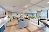 The Flex Lounge at 305 Winter St in Waltham, Massachusetts is a thoughtfully designed single-floor communal space that seamlessly blends comfort, functionality, and modern aesthetics. The open-plan layout is unified by wide-plank light hardwood flooring accented with inlaid geometric patterns, while a striking coffered ceiling with recessed lighting adds architectural depth and a sense of generous vertical space throughout.

The lounge area serves as the heart of the space, anchored by a large curved sectional sofa in warm beige, complemented by colorful throw pillows and a set of navy blue ottoman cubes. Nearby, a cozy seating nook features contemporary wingback and lounge chairs arranged around a low table, with built-in open shelving along the wall offering both display and storage. A large wall-mounted television with flanking speakers makes this corner ideal for media viewing and social gatherings.

Toward the perimeter, floor-to-ceiling windows flood the dining and café zone with abundant natural light and offer views of the surrounding landscape. This area features a long communal table surrounded by vibrant olive-green chairs, creating an inviting setting for collaborative work or casual dining. A well-appointed kitchenette with counter seating, coffee station, and cabinetry supports everyday use. The overall ambiance is bright, welcoming, and versatile — purpose-built for both productivity and relaxation.