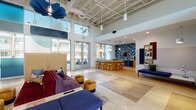 This single-story clubhouse in Mesa, Arizona delivers a vibrant, contemporary design defined by soaring ceilings with an exposed structural grid and expansive floor-to-ceiling windows that flood the interior with natural light and frame views of the surrounding courtyard and palm-lined grounds. Large-format tile flooring anchors the open main lounge, where bold, colorful furnishings—including a deep purple sofa, gold velvet ottomans, and navy seating—are arranged around a central partition, creating an inviting social hub. Distinctive blue floral pendant fixtures hang overhead, adding a sculptural, artistic accent to the space.

A curved natural wood bar with rattan stools serves as a focal point near the lounge, backed by a striking blue-and-teal chevron tile accent wall with an integrated television and cabinetry. Pendant lighting with warm-toned shades completes this stylish gathering area. Separate alcoves offer more intimate settings, featuring wood-paneled ceilings, patterned wallcoverings, a small bistro table, and lounge seating. An entry foyer showcases a diagonal wood slat screen partition, a built-in bench, and secure parcel lockers, combining practicality with thoughtful design. Glass doors throughout connect seamlessly to the outdoor courtyard.