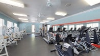 This fitness center offers a spacious, single-floor layout designed to accommodate a variety of exercise needs. The room features light blue walls accented by a bold coral stripe along the mirror line, creating an energetic yet approachable atmosphere. A large wall of mirrors runs along one side, visually expanding the space and allowing users to monitor their form during workouts.

The cardio section is well-stocked with multiple treadmills, elliptical machines, and stationary bikes arranged in organized rows. Strength and cable machines are positioned toward the back of the room, offering options for resistance and functional training. The dark rubber flooring provides a durable, cushioned surface suitable for both cardio and strength activities.

Overhead, flush-mounted fluorescent light fixtures provide consistent, bright illumination throughout, complemented by ceiling fans that promote air circulation. Natural light filters in through windows on multiple walls, enhancing the open feel of the space. Crown molding and a textured ceiling add a polished finish. The overall design balances functionality with a clean, welcoming aesthetic suited for regular fitness use.
