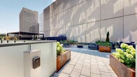 Located at 70 Nicholas St in Ottawa's dynamic urban core, this outdoor amenity terrace is a thoughtfully designed ground-level space that offers residents a well-appointed retreat from city life. The terrace is finished with large-format stone pavers throughout, creating a clean and cohesive aesthetic that ties together its various functional zones.

A generous open-air lounge area provides ample seating with modern Adirondack-style chairs arranged around planted garden beds featuring evergreen shrubs and seasonal plantings, offering both greenery and a sense of privacy. Nearby, a second seating zone is outfitted with contemporary benches and low coffee tables, ideal for relaxed gatherings with views of the surrounding cityscape.

The outdoor kitchen area is a standout feature, equipped with a stainless steel built-in gas grill, dedicated prep counter with granite surface, and a utility sink — all sheltered beneath a sleek metal pergola structure. A dark slatted metal privacy fence defines the perimeter, lending an architectural edge to the space.

Completing the terrace is a gravel-surfaced pet relief area, enclosed by matching dark metal fencing and integrated with young plantings. Together, these amenities create a versatile, well-landscaped outdoor environment suited for everyday relaxation and social gatherings alike.