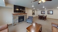 This single-floor community space is thoughtfully designed with an open-concept layout that seamlessly connects the kitchen, living, and dining areas into one cohesive and functional environment. The fully equipped kitchen is a standout feature, showcasing rich warm-toned wood cabinetry, light stone countertops, stainless steel appliances including a refrigerator, dishwasher, and range, and a central island with bar seating — all set atop durable light hardwood-style flooring. Recessed lighting and pendant fixtures keep the kitchen bright and inviting. The living area is anchored by a striking floor-to-ceiling stacked stone fireplace topped with a wall-mounted flat-screen television, creating a cozy focal point for gatherings. Plush neutral carpeting, comfortable leather sofas and accent chairs, and a ceiling fan contribute to the warm and relaxed ambiance. Large windows flood the space with natural light while offering views of the surrounding exterior. A dedicated dining area with a dark wood table and ample seating rounds out the space, making it well-suited for both casual get-togethers and organized events. The overall design balances comfort and practicality, making this a versatile and welcoming communal destination.