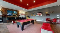 Located in Westerville, Ohio, this single-story community clubhouse is designed with bold, eclectic style and a wide range of amenities. The welcoming entryway features hardwood-style flooring, a patterned crimson area rug, and a marquee-lit theater entrance that sets a lively tone throughout.

The expansive game room is a standout, boasting a striking red lacquered tray ceiling with recessed lighting, a full-size billiard table, skeeball lane, and a built-in bar area with stone accent walls, bar seating, and multiple flat-screen TVs. Decorative elements including antler mounts, a Union Jack artwork, and retro signage give the space a distinctive personality.

Adjacent is a private screening room styled after a classic theater, with dark walls, vintage movie posters, plush leather seating, and a projection system — ideal for intimate screenings or gatherings.

The refreshment station features dark wood cabinetry, granite countertops, a commercial beverage dispenser, ice machine, and Starbucks offerings. A reading lounge offers a sophisticated retreat with a dramatic diamond-grid bookshelf, red leather armchairs, and natural light from large windows.

The fitness center is generously sized with high ceilings, large windows, ceiling fans, a full array of strength machines, cardio equipment, and mirrored walls for an open, airy workout environment.