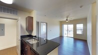 This single-floor residence offers a thoughtfully designed open-concept layout that seamlessly connects the kitchen and living areas, creating a spacious and inviting atmosphere. The kitchen is a standout feature, showcasing rich dark wood cabinetry, sleek black granite countertops with a double stainless steel sink, and a breakfast bar that opens directly into the main living space — ideal for both everyday living and entertaining. The living area is finished with warm hardwood flooring that extends throughout, complemented by soft, neutral-toned walls that enhance the bright and airy feel of the space. A ceiling fan overhead adds both style and functional comfort. Large sliding glass doors flood the living area with abundant natural light and provide direct access to a private balcony with open outdoor views. A doorway visible from the main living space leads to an additional room, also benefiting from natural light through its own window. The overall ambiance of this residence is clean, modern, and welcoming, with a cohesive design that balances practicality with contemporary style.