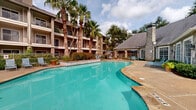 Located at 2742 Jeanetta Rd in Houston, Texas, this outdoor amenity area serves as a central courtyard and recreational hub. The centerpiece is a generously sized freeform swimming pool with vivid turquoise water, framed by a wide concrete pool deck furnished with lounge chairs and shaded seating areas ideal for relaxation. Tall, mature palm trees are planted throughout the courtyard, lending a tropical ambiance that complements the warm Houston climate.

Adjacent to the pool is a stone-accented clubhouse structure with a pitched, shingled roof and large grid-pane windows. The building is accented by a prominent stone chimney and covered patio space, adding architectural character to the setting. Outdoor dining tables with blue market umbrellas provide a comfortable gathering space on the sun-drenched deck.

The courtyard is enclosed by a multi-story residential building with stucco and stone exterior finishes, featuring private balconies on each level that overlook the pool area. Wrought iron fencing borders the pool perimeter, while neatly trimmed hedges, ornamental plantings, and potted flowers enhance the landscaped surroundings. The open sky above and abundant natural light create a bright, resort-style atmosphere throughout the outdoor space.