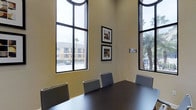 The conference room at 1935 Northstar Way in San Marcos is a professionally designed meeting space that combines functionality with an open, airy atmosphere. The room is immediately distinguished by its dramatically high ceilings and expansive floor-to-ceiling windows framed in dark metal, which flood the interior with abundant natural light and offer views of the surrounding palm-lined outdoor environment. The large-pane windows are arched at the top, adding an architectural elegance to the space. At the center of the room sits a substantial dark-finished conference table surrounded by a set of coordinating gray ergonomic chairs, providing comfortable seating for meetings and collaborative sessions. The walls are finished in a warm neutral tone, contributing to a calm and focused atmosphere. Two framed art pieces are displayed on the primary wall, adding a polished, professional touch to the décor. A room scheduling panel is mounted near the entrance, offering convenient access management. The combination of soaring ceilings, generous glazing, and thoughtful furnishings makes this conference room a practical and inviting venue for professional gatherings, presentations, or collaborative work sessions.