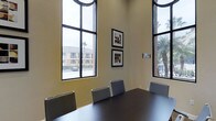 The conference room at 1935 Northstar Way in San Marcos is a professionally designed meeting space that combines functionality with an open, airy atmosphere. The room is immediately distinguished by its dramatically high ceilings and expansive floor-to-ceiling windows framed in dark metal, which flood the interior with abundant natural light and offer views of the surrounding palm-lined outdoor environment. The large-pane windows are arched at the top, adding an architectural elegance to the space. At the center of the room sits a substantial dark-finished conference table surrounded by a set of coordinating gray ergonomic chairs, providing comfortable seating for meetings and collaborative sessions. The walls are finished in a warm neutral tone, contributing to a calm and focused atmosphere. Two framed art pieces are displayed on the primary wall, adding a polished, professional touch to the décor. A room scheduling panel is mounted near the entrance, offering convenient access management. The combination of soaring ceilings, generous glazing, and thoughtful furnishings makes this conference room a practical and inviting venue for professional gatherings, presentations, or collaborative work sessions.