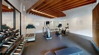 Located at 2150 Boulevard De Maisonneuve Ouest in Montreal, this well-appointed fitness facility offers a thoughtfully designed workout space within a compact yet efficiently utilized floor plan. The gym's most striking architectural feature is its striking suspended ceiling of warm-toned wood slats, accented by integrated linear LED lighting that creates a modern, inviting ambiance throughout the space.

The fitness area is equipped with a comprehensive selection of Matrix-brand cardio machines, including treadmills, elliptical trainers, upright stationary bikes, and spinning cycles, providing a full range of aerobic training options. A dedicated free weights section includes a dumbbell rack, while a cable machine and adjustable weight bench support strength training workouts.

A decorative vertical wood slat partition divides the space with visual interest while maintaining an open feel. Floor-to-ceiling mirrors along one wall amplify the sense of space and allow users to monitor their form during exercise. Durable rubber flooring covers the entire workout area, ensuring safety and comfort underfoot.

Natural light filters in through large windows that offer views of the surrounding streetscape, complemented by a wall-mounted climate control unit. Cylinder pendant spotlights provide additional targeted illumination. The clean white walls and bright, airy atmosphere make this gym a functional and aesthetically refined facility.