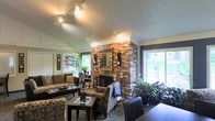 This single-floor space exudes a cozy yet characterful ambiance, anchored by a dramatic floor-to-ceiling brick fireplace that serves as the focal point of the main living area. The fireplace surround, constructed from rich, warm-toned brick, adds a timeless architectural element and a sense of solidity to the room. Vaulted ceilings rise above the living space, enhancing the open, airy feel and accommodating a sleek track lighting system that provides adjustable illumination throughout. Large windows on multiple walls flood the interior with generous natural light, offering views of lush greenery outside and further connecting the interior to the outdoors. The living area is thoughtfully arranged with comfortable seating including sofas and patterned armchairs, complemented by wood-framed coffee tables that add warmth and functionality. Decorative touches such as wall art, candle sconces flanking the fireplace, and potted plants bring life and personality to the space. The textured ceiling adds depth and character, while the overall layout promotes a welcoming, lived-in atmosphere ideal for both relaxing and entertaining. This is a well-appointed space that balances rustic charm with comfortable, everyday livability.