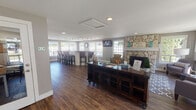 Located at 4710 S 302nd Ln in Auburn, WA, this single-floor community or clubhouse-style space combines a warm lakeside aesthetic with functional, well-appointed areas designed for comfort and utility. Rich hardwood-style flooring runs throughout, anchoring the cohesive design language of the interior.

The central lounge area serves as the heart of the space, highlighted by a striking floor-to-ceiling stone fireplace with a white mantel, decorated with a "Welcome to the Lake" sign and teal glass accents. Large multi-pane windows surround the room, inviting generous natural light and offering views of the surrounding landscape. The lounge is furnished with a mix of upholstered seating, a patterned area rug, and a flat-screen TV mounted on the wall.

Adjacent to the lounge is a curved bar-height counter with granite-style surfaces and padded bar stools, illuminated by an overhead track lighting system. This area is lined with expansive windows that further enhance the bright, airy atmosphere.

A transitional hallway connects to a private office or administrative area, featuring framed artwork along the gray-painted walls, a glass-paneled door, and a small workstation. A separate entrance with sidelight windows and exit signage provides clear access and egress. The overall ambiance is inviting and polished, blending casual lakeside charm with practical community-oriented functionality.