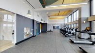 This single-floor fitness facility offers a thoughtfully designed workout environment that combines functionality with contemporary architectural detail. The space is immediately distinguished by its elevated ceilings, which incorporate a recessed section finished with warm wood paneling and decorative acoustic tiling in contrasting green and gold tones, adding visual interest overhead. A row of large industrial-style pendant lights in muted grey tones lines the length of the main floor, providing consistent, well-distributed illumination throughout.

The main workout area features a generous row of cardio equipment, including elliptical trainers and stationary bikes, positioned along a wall of large windows that allow ample natural light to enter the space. The flooring consists of durable, speckled rubber or composite gym tiles, practical for high-traffic athletic use. The far end of the space opens toward an additional exercise or equipment area, visible through a mirrored partition.

Accent walls in deep charcoal grey contrast with the crisp white upper walls, while teal and yellow door and trim details introduce pops of color. A recessed water fountain station is built into one wall, adding a convenient, integrated amenity to the facility's clean, organized layout.
