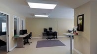 This single-floor office space is designed with practicality and functionality in mind, offering an open and organized work environment. The main workspace area features multiple computer workstations, each equipped with desktop monitors, keyboards, and ergonomic office chairs, making it well-suited for collaborative or individual work tasks. A printer and additional office equipment are stationed along the back wall, ensuring essential resources are easily accessible to occupants.

The interior is finished with neutral beige walls that create a calm and professional atmosphere, complemented by light-toned flooring throughout. Large recessed fluorescent panel lights are mounted in the ceiling, providing bright, even illumination across the workspace. A low-pile area rug beneath one of the workstations adds a subtle touch of comfort and helps define the space.

To one side, a reception or check-in counter is visible, suggesting the space may serve a client-facing function in addition to standard office operations. Windows along the perimeter allow for some natural light to filter into the room. Overall, the layout is clean and efficiently organized, prioritizing workflow and ease of movement, making it a practical setting for a variety of professional or administrative purposes.