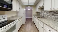 This two-bedroom, one-bathroom unit is thoughtfully laid out across a single floor, offering a well-organized and functional living space. The floor plan reveals a logical flow between living, dining, kitchen, and bedroom areas, all connected by consistent design elements and neutral color palettes.

The living and dining areas feature dark hardwood-style flooring that extends throughout the main spaces, complemented by neutral gray walls and white baseboards. Large picture windows in the living area allow for generous natural light and exterior views, with curtain panels providing an option for privacy. A pendant light fixture anchors the dining area, which opens directly into the kitchen.

The galley-style kitchen is well-appointed with white cabinetry, granite-look countertops, a tile mosaic backsplash, a stainless steel sink, and white appliances including a refrigerator, range, and dishwasher. Overhead lighting illuminates the full length of the workspace.

Both bedrooms are carpeted with large windows that bring in natural light. The entry hallway features carpet flooring and a solid wood interior door. The bathroom includes a full tub and shower combination with white tile surround, a dual-function showerhead, toilet, and vanity set on neutral tile flooring.