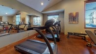 This dedicated fitness room offers a comprehensive workout environment thoughtfully designed for both cardio and strength training. The space features vaulted ceilings that lend an open, airy feel, complemented by recessed lighting and ceiling fans that help maintain a comfortable atmosphere during exercise. Rich, polished concrete flooring with warm tones runs throughout, providing a durable and easy-to-maintain surface.

The cardio zone is well-appointed with multiple treadmills and stationary bikes, while a full-length mirror along one wall enhances the sense of space and allows for proper form checks. A wall-mounted flat-screen television adds entertainment options for longer workout sessions. The strength training area features a multi-station cable machine, an adjustable weight bench, and a full dumbbell rack, providing everything needed for a thorough resistance workout.

Natural light filters in through wood-framed windows with blinds, offering views to the outdoors while maintaining privacy and light control. Warm-toned artwork on the walls adds a welcoming touch to the functional space. A coat hook rail and waste bin reflect thoughtful attention to everyday convenience. The room connects to an adjacent area visible through a large mirror pass-through, suggesting seamless integration with the broader facility.