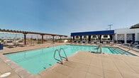 Located at 3675 T St in Sacramento's Midtown neighborhood, this rooftop amenity deck offers an expansive outdoor retreat with sweeping views of the Sacramento skyline. At its center is a large, sparkling swimming pool equipped with stainless steel entry rails and an accessibility lift, surrounded by generous sun-drenched pool decking finished in large-format concrete tiles. Wood-beamed pergolas line the perimeter, providing shaded lounging areas furnished with chaise lounges and seating for relaxation throughout the day. A bold blue canopy structure anchors one side of the deck, housing an entertainment and bar area complete with mounted televisions and additional seating. An outdoor kitchen and dining area with a tiled accent wall adds to the entertaining options, while private cabana spaces with curtained enclosures and wicker furniture offer a more secluded experience. A tiled outdoor shower, featuring a striking navy and white hexagonal tile design, provides convenience for pool users. Glass wind barriers along the deck's edge preserve the open, airy atmosphere while maintaining unobstructed views of the surrounding cityscape. The overall ambiance is modern and resort-inspired, ideal for leisure and social gatherings under the abundant Sacramento sun.