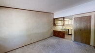 Located at 2202 Brunswick Ct in Morgantown, West Virginia, this compact single-floor unit offers an efficient and functional layout comprising a kitchen and a full bathroom. The main living area features neutral-toned carpet flooring, textured walls, and a textured ceiling, all framed by wood crown molding. A large window near the entry door allows natural light to filter in and provides a view of the surrounding greenery, while a wall-mounted HVAC unit ensures year-round climate control. The bold black entry door adds a contrasting design element to the otherwise neutral interior palette.

The kitchen area transitions from the carpeted living space to hard-surface wood-look flooring and is equipped with a freestanding white gas range, a double-basin sink, and a built-in dishwasher. A wood-toned kitchen island with lower cabinet storage provides additional workspace and visual separation between the kitchen and living areas. Open wood shelving mounted above creates additional storage and an open, airy feel in the kitchen zone.

A dedicated storage area features a built-in wood dresser unit and an adjacent sliding wood-panel closet door, offering practical organizational options. The bathroom includes a clean, white tub-shower enclosure with chrome fixtures and hardware. Throughout the unit, warm wood tones in the cabinetry and trim provide cohesive character.
