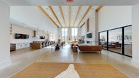 This single-floor community clubhouse showcases a striking blend of modern and rustic design elements throughout its expansive, open layout. The centerpiece of the space is a grand great room with dramatically high ceilings accented by exposed natural wood beams and polished concrete floors that reflect abundant natural light. Floor-to-ceiling black-framed windows line the far wall, offering sweeping views of the surrounding community and flooding the interior with daylight. A large stone fireplace anchors one side of the room, complete with a wall-mounted TV and a linear gas fire feature, while a generous seating area with cognac leather sofas and a glass coffee table creates a sophisticated lounge environment. A dedicated dining area with dark round tables and mixed seating provides ample space for group gatherings. The fully equipped kitchen features crisp white cabinetry with matte black hardware, a gas cooktop, stainless steel appliances, a deep utility sink, and open wood shelving — ideal for catered events or communal use. The entry lobby impresses with a curved ceiling detail, a statement chandelier, and polished concrete floors leading to a reception area framed by tall windows. Additional lounge nooks with minimalist furnishings and curated western-themed décor complete the thoughtfully designed space.