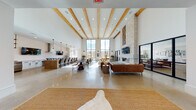 This single-floor community clubhouse showcases a striking blend of modern and rustic design elements throughout its expansive, open layout. The centerpiece of the space is a grand great room with dramatically high ceilings accented by exposed natural wood beams and polished concrete floors that reflect abundant natural light. Floor-to-ceiling black-framed windows line the far wall, offering sweeping views of the surrounding community and flooding the interior with daylight. A large stone fireplace anchors one side of the room, complete with a wall-mounted TV and a linear gas fire feature, while a generous seating area with cognac leather sofas and a glass coffee table creates a sophisticated lounge environment. A dedicated dining area with dark round tables and mixed seating provides ample space for group gatherings. The fully equipped kitchen features crisp white cabinetry with matte black hardware, a gas cooktop, stainless steel appliances, a deep utility sink, and open wood shelving — ideal for catered events or communal use. The entry lobby impresses with a curved ceiling detail, a statement chandelier, and polished concrete floors leading to a reception area framed by tall windows. Additional lounge nooks with minimalist furnishings and curated western-themed décor complete the thoughtfully designed space.