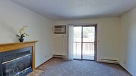 This single-floor space presents a practical and comfortable layout designed for everyday living. The main living area is carpeted and features a classic fireplace with a wood-trimmed mantel and tile surround, serving as a natural focal point of the room. A sliding glass door floods the space with natural light and opens directly onto a private balcony, offering a pleasant outdoor view with glimpses of surrounding trees. A wall-mounted air conditioning unit provides climate control, complemented by baseboard heating along the perimeter walls for year-round comfort. The textured ceiling adds a traditional character to the interior. The neutral wall tones and understated finishes create a clean, versatile backdrop well-suited to a variety of furnishing styles. Overall, the space balances functional amenities with a relaxed, approachable ambiance that makes it easy to personalize and settle into.