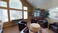 This single-floor space exudes a refined and comfortable ambiance, thoughtfully designed to balance elegance with warmth. The centerpiece of the main living area is a dramatic vaulted ceiling that soars overhead, amplifying the sense of openness and grandeur. A striking arched window, framed with decorative leaded glass detailing, anchors one wall and floods the room with natural light, complemented by additional wide windows fitted with horizontal blinds for adjustable privacy. The warm, neutral-toned walls provide a soothing backdrop throughout. The living area is appointed with plush dark leather armchairs arranged around a soft, round upholstered ottoman, creating an intimate and inviting seating arrangement atop a textured area rug. A stately dark wood display cabinet with glass-paneled doors serves as a prominent focal point, offering both storage and decorative display space, and is accented by lush greenery and decorative accessories. Soft carpeted flooring runs throughout, adding to the cozy atmosphere. Warm ambient lighting from a classic floor lamp enhances the space during evening hours. Overall, the space offers a harmonious blend of architectural character and comfortable furnishings, making it well-suited for relaxed gatherings or quiet retreat.