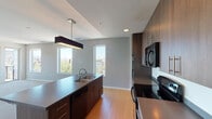 This single-floor unit presents a well-designed contemporary layout with distinct living, kitchen, and bedroom areas. The kitchen is a standout feature, equipped with dark wood-grain cabinetry extending to the ceiling, gray composite countertops, black appliances including an electric range, built-in microwave, and a stainless steel refrigerator. A central kitchen island with a double stainless steel sink and pendant lighting adds both functionality and style, while light hardwood flooring ties the space together.

The open living area features soft carpet flooring and is bathed in natural light through large sliding glass doors that open to an outdoor balcony, offering views of the surrounding cityscape. A separate carpeted bedroom provides additional city views through a bright, sun-filled window.

A dedicated utility area houses a stacked washer and dryer alongside a water heater and HVAC system. The bathroom is finished with tile flooring, a wide dual-sink vanity with wood-grain cabinetry, a large mirror, and warm overhead lighting. Throughout the unit, a neutral color palette and clean architectural lines create a cohesive, modern ambiance suited for urban living.