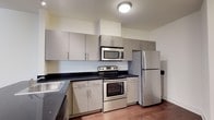 This single-floor space showcases a well-appointed kitchen designed for both functionality and style. The kitchen features a full suite of stainless steel appliances, including a refrigerator, electric range, and over-the-range microwave, offering a cohesive and contemporary aesthetic. Dark stone countertops provide a striking contrast against the light grey, flat-front cabinetry that lines the walls, offering generous storage both above and below the counter. The lower cabinets extend along an L-shaped layout, maximizing workspace efficiency. A stainless steel sink is positioned along one counter run, complementing the overall metallic finish throughout. The warm-toned hardwood flooring adds a natural element that balances the cool tones of the cabinetry and appliances. Walls are finished in a neutral off-white tone, contributing to a clean, bright atmosphere. Overhead, a flush-mount ceiling light provides practical illumination across the workspace. The kitchen's layout is thoughtfully arranged to ensure ease of movement and accessibility, making it a practical and visually appealing central feature of the space.