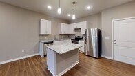 Located at 15450 Ruggles St in Omaha, Nebraska, this single-floor residence offers a well-designed layout with modern finishes throughout. The kitchen serves as a functional centerpiece, featuring crisp white shaker cabinetry, granite countertops with subtle veining, a central island with pendant lighting overhead, and a full suite of stainless steel appliances including a refrigerator, electric range, and dishwasher. Rich hardwood-style flooring runs through the kitchen and connects seamlessly to the open living area, which is bathed in natural light through multiple windows offering views of the surrounding exterior. Recessed lighting throughout the main living space ensures bright, even illumination. The bedroom offers plush carpeted flooring, a dedicated window for natural light, and a ceiling light fixture, creating a comfortable and quiet retreat. The bathroom is clean and well-appointed, featuring a combination tub and shower with a curved curtain rod, a toilet, and a vanity with dark countertops and ample drawer storage, all set on wood-style flooring with neutral taupe walls tying the space together. The overall aesthetic is contemporary and cohesive, with warm neutral tones and quality finishes evident throughout every room.