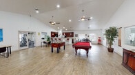 This single-floor space is designed as a versatile community or recreational facility, offering a well-rounded mix of leisure, dining, and meeting areas. The centerpiece of the space is a generously sized open game room with soaring vaulted ceilings, light hardwood-style flooring, and dual ceiling fans that enhance the airy atmosphere. The room is anchored by a full-size red felt pool table and a foosball table, providing ample entertainment options. Large windows along the perimeter allow natural light to fill the space and offer views of the surrounding exterior.

Adjacent to the game room is a fully equipped kitchen featuring rich dark wood cabinetry, granite-style countertops, stainless steel appliances including a gas range and refrigerator, and tile flooring. A raised breakfast bar with seating connects the kitchen to the main area, making it functional for communal gatherings. A dining area with high-top tables and chairs is positioned nearby, ideal for casual meals or social interaction.

A dedicated conference room offers a more focused environment, furnished with a long dark conference table, modern seating, a wall-mounted flat-screen TV, and a full bank of windows that flood the room with natural light. The overall design is clean, bright, and functional, catering to both social and professional needs.