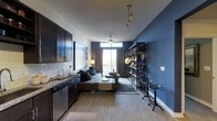 This thoughtfully designed single-floor space blends modern aesthetics with functional living in an open-concept layout. The kitchen is outfitted with rich dark cabinetry, granite countertops, a subway tile backsplash, and stainless steel appliances, offering both style and practicality. The living area flows seamlessly from the kitchen and is anchored by bold navy blue accent walls that lend a sophisticated, contemporary feel to the space. Light wood-look flooring runs throughout, adding warmth and continuity. A ceiling fan with integrated lighting serves the central living zone, while a decorative track light fixture adds an industrial touch overhead. An open metal shelving unit with a ladder-style design provides both storage and a striking visual element, complemented by matching industrial-style bar stools. The space is bathed in natural light from a large sliding glass door that opens to a private balcony, framed by floor-to-ceiling drape panels. The living area comfortably accommodates a sectional sofa and coffee table arrangement, making it ideal for relaxing or entertaining. The overall ambiance is modern and curated, with a carefully balanced mix of dark, moody tones and natural light creating an inviting, upscale atmosphere.