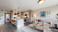 This thoughtfully designed single-floor residence combines modern aesthetics with functional living across several well-appointed spaces. The open-concept kitchen and living area serves as the heart of the unit, where light wood cabinetry, white quartz countertops, and a subway tile backsplash complement stainless steel appliances, including a gas range and built-in microwave. A central island with seating for multiple provides both casual dining and workspace, illuminated by striking matte black pendant lights overhead. An in-unit stacked washer and dryer is conveniently tucked nearby. The living area flows naturally from the kitchen, featuring large windows and a sliding glass door that invite natural light and connect to an outdoor space. Dark hardwood-style flooring runs throughout the main areas, grounding the neutral, warm palette. The bedroom offers tree-lined window views and exposed concrete accent details, lending an urban-industrial character. A versatile flex room with a large window and plush carpet provides an ideal home office or secondary bedroom. Two bathrooms include one full bath featuring a patterned tile tub surround and a floating vanity with matte black fixtures, and a second with tile flooring and a natural light window. The overall ambiance is contemporary, calm, and cohesive.