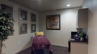 This single-level wellness treatment room offers a calm and professional environment designed for therapeutic services such as massage or bodywork. The centerpiece of the space is a padded treatment table draped in a rich purple covering and accented with a yellow decorative pillow, providing a welcoming and comfortable focal point for clients.

The walls are finished in soft, neutral tones and adorned with a curated gallery of framed artwork, including a large mosaic-style piece in warm earth tones and a symmetrical arrangement of six smaller framed prints. A tall decorative ficus tree adds a natural, organic element to the room, enhancing the overall sense of tranquility.

A dedicated utility station along one wall features light gray cabinetry — both upper and lower — with a dark countertop and a stainless steel sink, providing a practical and clean workspace for the practitioner. Recessed lighting illuminates the room with a soft, even glow, while patterned carpet adds texture underfoot.

Additional furnishings include a practitioner's stool and a portable massage chair, indicating versatility in the types of treatments offered. The space opens to a hallway, suggesting it is part of a larger professional wellness or medical facility. Overall, the room balances functionality with a soothing aesthetic.