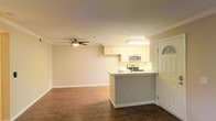 This single-floor residence offers an open and functional layout that seamlessly connects the kitchen and living areas. The kitchen is equipped with stainless steel appliances, including a built-in microwave and a range, complemented by light-toned cabinetry that provides ample storage. A peninsula-style breakfast bar separates the kitchen from the living space, offering both a casual dining option and an efficient use of space. Under-cabinet lighting brightens the workspace, enhancing the kitchen's practical appeal. The living area is generously proportioned, featuring rich hardwood flooring that adds warmth and character throughout. Crown molding lines the ceiling, lending a refined architectural detail to the space. A ceiling fan with an integrated light fixture is centrally positioned, promoting air circulation and adding to the room's comfort. The entry door, featuring a decorative half-moon transom window, allows natural light to filter into the foyer. Neutral wall tones throughout create a clean, versatile backdrop well-suited to a variety of interior styles. The overall ambiance is bright, open, and move-in ready, reflecting a well-maintained and thoughtfully designed living environment.